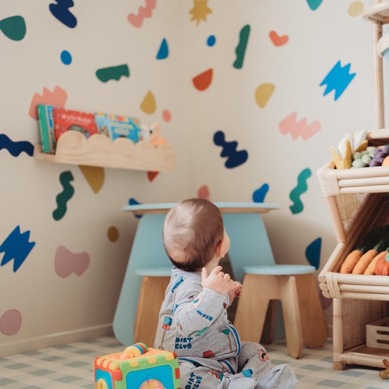 May include: A child's playroom with a light blue table and stools, a wooden shelf with books, and a multi-colored toy. The walls are decorated with colorful abstract shapes. A wooden shelving unit displays toy vegetables.