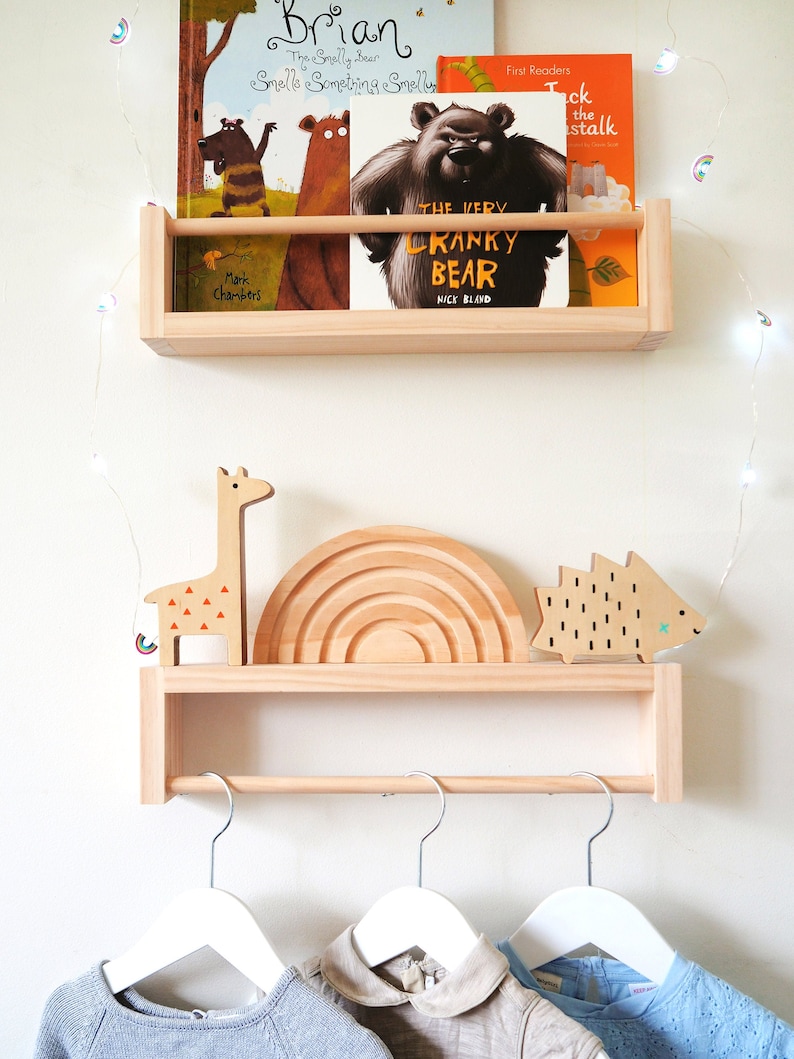 May include: A wooden shelf with a rail for hanging clothes. The shelf has a top section for displaying books and toys. The books include "Brian: The Smelly Bear Smells Something Smelly" and "The Very Cranky Bear". The toys include a wooden giraffe, a rainbow, and a hedgehog.