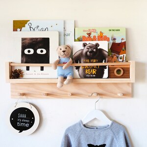 May include: A wooden wall-mounted shelf with hooks, featuring a small stuffed bear, a round sign that says "SHHH... it's sleep time", and a variety of children's books.