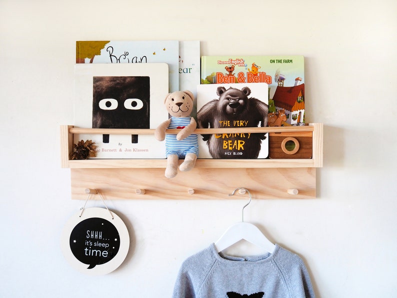 May include: A wooden wall-mounted shelf with hooks, featuring a small stuffed bear, a round sign that says "SHHH... it's sleep time", and a variety of children's books.