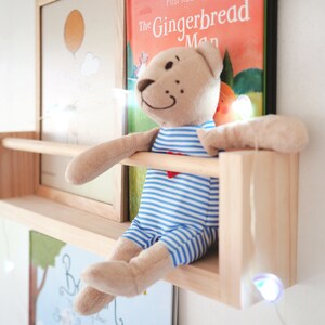May include: A wooden shelf with a stuffed teddy bear wearing a blue and white striped shirt sitting on it. The shelf is mounted on a white wall with a string of fairy lights hanging down. The shelf has a small wooden bar across the front. There are two framed pictures behind the shelf, one with a yellow sun and the other with the text "The Gingerbread Man".