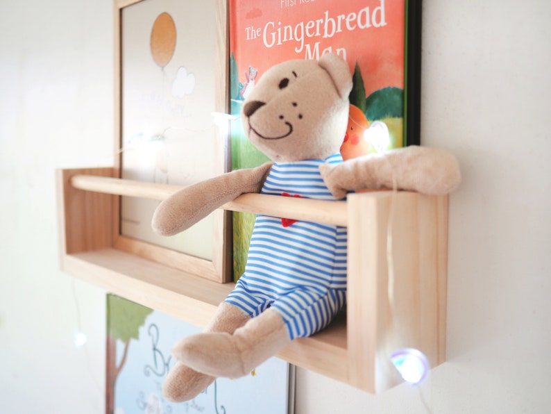 May include: A wooden shelf with a stuffed teddy bear wearing a blue and white striped shirt sitting on it. The shelf is mounted on a white wall with a string of fairy lights hanging down. The shelf has a small wooden bar across the front. There are two framed pictures behind the shelf, one with a yellow sun and the other with the text "The Gingerbread Man".