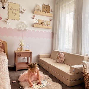 May include: A child's bedroom with a beige modular sofa, a pink side table, and decorative shelves. The walls are painted white and pink with a cloud-like border. A young girl is playing with a wooden puzzle on a fluffy rug.