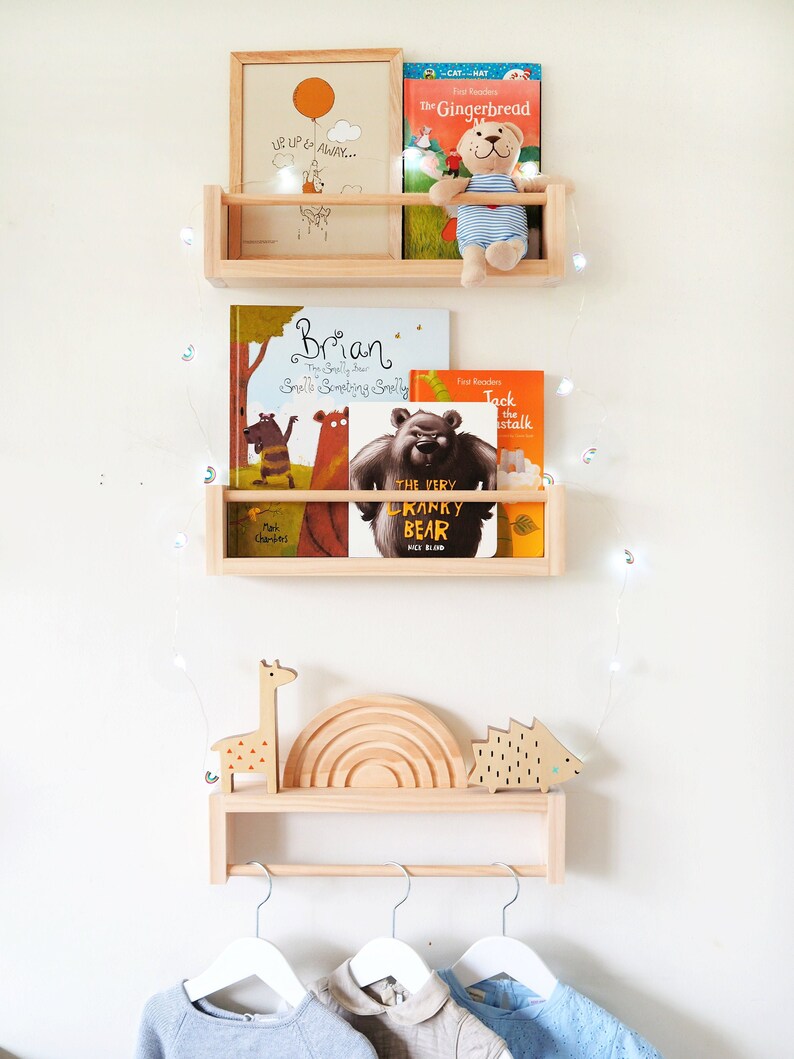 May include: Three wooden shelves with a white background. The top shelf has a framed print with the text "UP UP & AWAY..." and a stuffed animal. The middle shelf has three children's books, including "Brian The Snelly Bur" and "The Very Cranky Bear". The bottom shelf has a wooden giraffe, rainbow, and hedgehog, and a wooden rod with three white hangers.