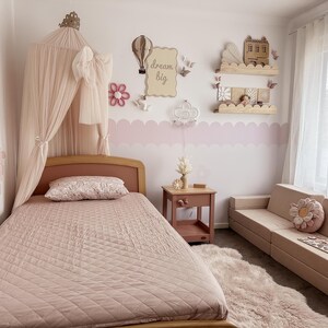 May include: A light pink bedroom with a bed, nightstand, and sofa. The bed has a pink quilted comforter and a canopy. The wall features a "dream big" sign, decorative shelves, and a pink scalloped border. A fluffy rug and flower pillows add to the decor.