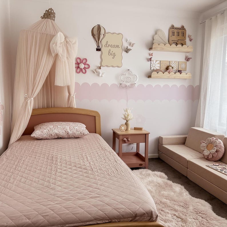 May include: A light pink bedroom with a bed, nightstand, and sofa. The bed has a pink quilted comforter and a canopy. The wall features a "dream big" sign, decorative shelves, and a pink scalloped border. A fluffy rug and flower pillows add to the decor.