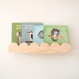 May include: A wooden cloud-shaped shelf holds four children's books with animal illustrations. The books have green, blue, and light green covers. The shelf is made of light-colored wood and is mounted on a white wall.