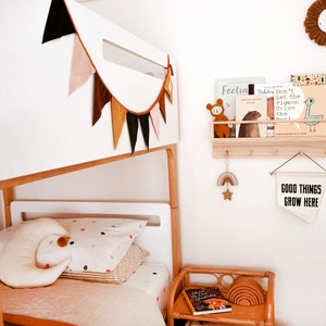 May include: A white bunk bed with a colorful fabric bunting hanging above it. The bed has a white mattress and a white headboard. There is a small wooden shelf with books on it above the bed. The shelf is attached to the wall. There is a small wooden table with a book and a set of rainbow-colored wooden rings on it. There is a white sign hanging on the wall that says "Good Things Grow Here".