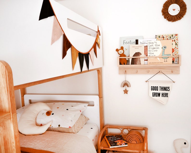 May include: A white bunk bed with a colorful fabric bunting hanging above it. The bed has a white mattress and a white headboard. There is a small wooden shelf with books on it above the bed. The shelf is attached to the wall. There is a small wooden table with a book and a set of rainbow-colored wooden rings on it. There is a white sign hanging on the wall that says "Good Things Grow Here".