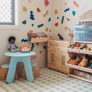 May include: A child's playroom with a light blue table and stools, a doll, and a toy phone. The room features a wooden play kitchen, a bookshelf, and a display of toy fruits and vegetables. Colorful shapes decorate the walls, and the floor has a checkered pattern.