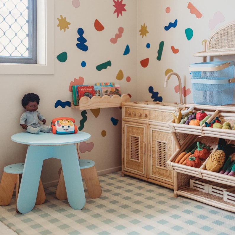 May include: A child's playroom with a light blue table and stools, a doll, and a toy phone. The room features a wooden play kitchen, a bookshelf, and a display of toy fruits and vegetables. Colorful shapes decorate the walls, and the floor has a checkered pattern.