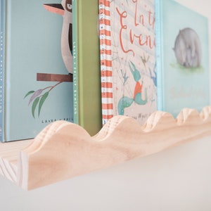 Peut inclure: Une &eacute;tag&egrave;re en bois avec un bord festonn&eacute;, tenant une rang&eacute;e de livres pour enfants avec des illustrations color&eacute;es. Les livres sont intitul&eacute;s "In the Evening" et "Laura & Philip Bunting".