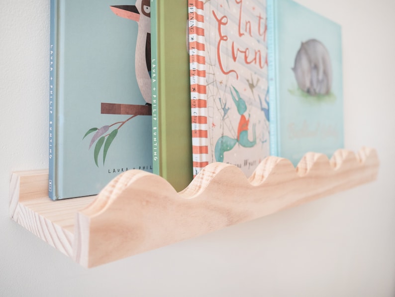 Peut inclure: Une &eacute;tag&egrave;re en bois avec un bord festonn&eacute;, tenant une rang&eacute;e de livres pour enfants avec des illustrations color&eacute;es. Les livres sont intitul&eacute;s "In the Evening" et "Laura & Philip Bunting".