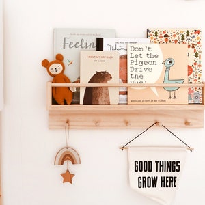 May include: A wooden shelf with hooks, holding a collection of children's books, a stuffed animal, and a banner that reads "GOOD THINGS GROW HERE".