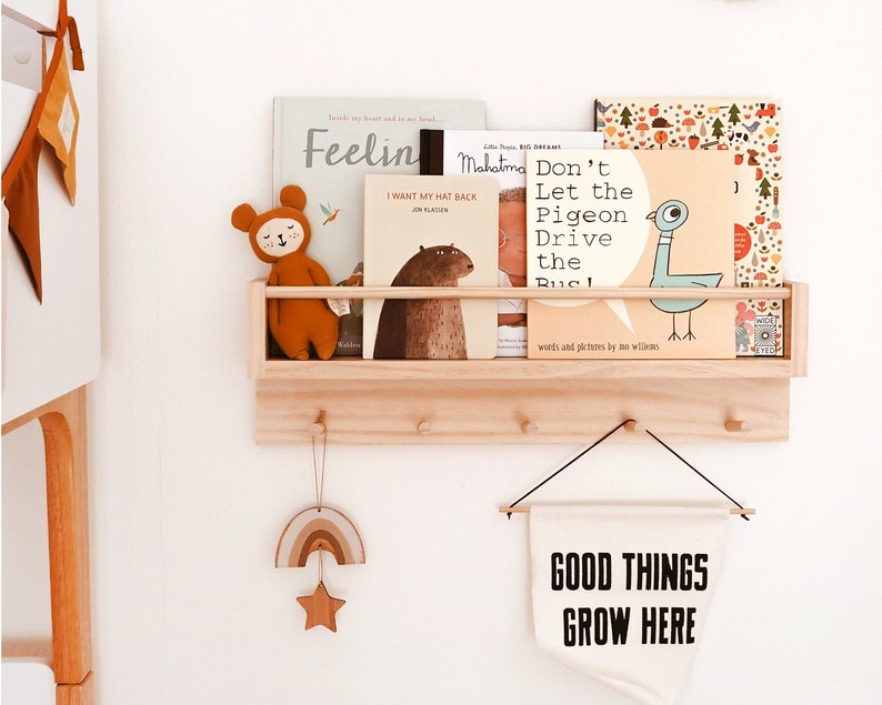 May include: A wooden shelf with hooks, holding a collection of children's books, a stuffed animal, and a banner that reads "GOOD THINGS GROW HERE".