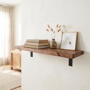 Può includere: Una mensola rettangolare in legno montata con due staffe in metallo nero. Sulla mensola ci sono due libri impilati, uno intitolato Essays in Texture, un piccolo vaso in ceramica marrone con steli d'erba essiccati e una stampa incorniciata.