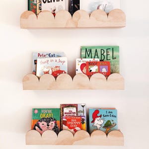 May include: Three wooden cloud-shaped shelves mounted on a white wall, each holding children's books. The books have colorful covers with titles like "Pete the Cat," "Mabel," and "I Am Sam-I-Am." The shelves are made of light-colored wood.