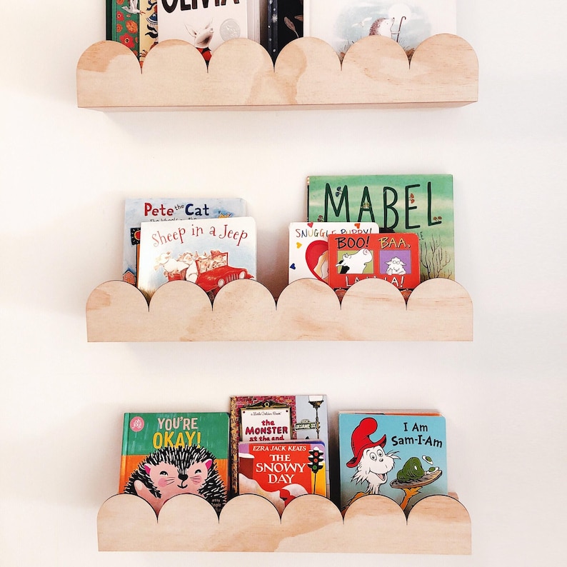 May include: Three wooden cloud-shaped shelves mounted on a white wall, each holding children's books. The books have colorful covers with titles like "Pete the Cat," "Mabel," and "I Am Sam-I-Am." The shelves are made of light-colored wood.