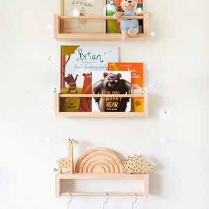 May include: A wooden shelf unit with three shelves, featuring a variety of children's books, a stuffed animal, and a picture frame. The top shelf has a picture frame with the text "Up, Up & Away..." and a stuffed animal bear. The middle shelf has three books, one titled "Brian", another titled "The Very Cranky Bear", and a third titled "Jack the Stalk". The bottom shelf has a wooden giraffe, a rainbow-shaped wooden toy, and a wooden hedgehog. The shelf unit is mounted on a white wall with a string of fairy lights.