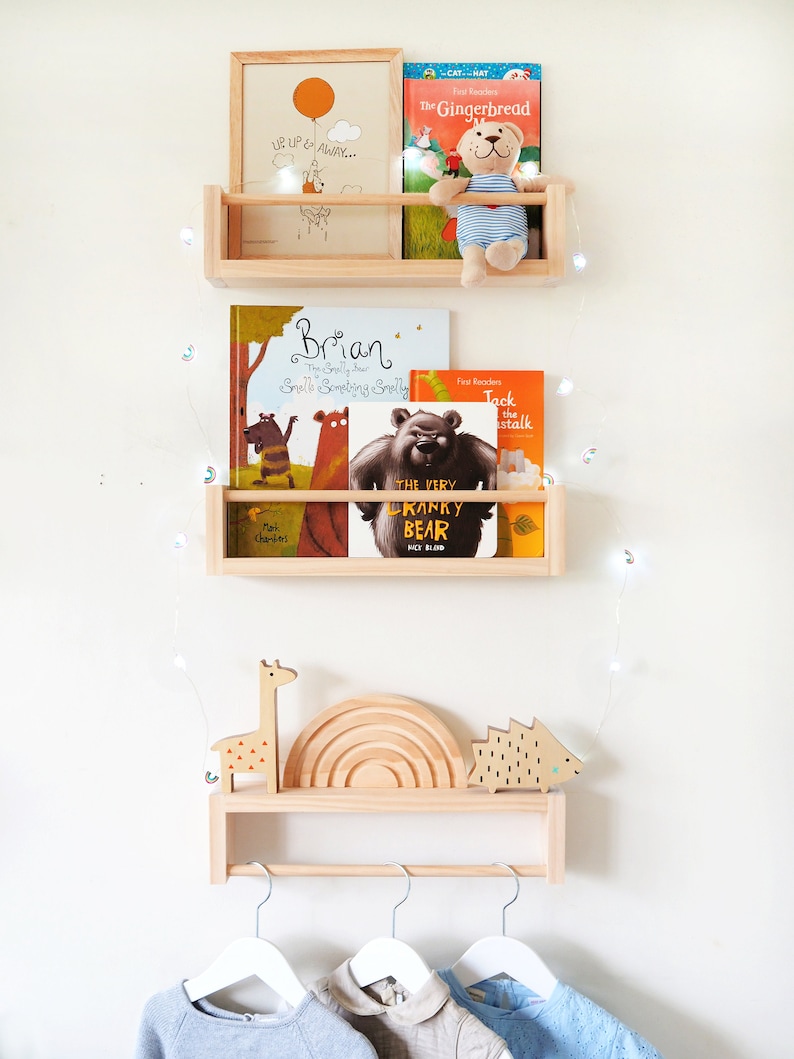 May include: A wooden shelf unit with three shelves, featuring a variety of children's books, a stuffed animal, and a picture frame. The top shelf has a picture frame with the text "Up, Up & Away..." and a stuffed animal bear. The middle shelf has three books, one titled "Brian", another titled "The Very Cranky Bear", and a third titled "Jack the Stalk". The bottom shelf has a wooden giraffe, a rainbow-shaped wooden toy, and a wooden hedgehog. The shelf unit is mounted on a white wall with a string of fairy lights.