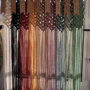 May include: A row of ten macrame plant hangers in various colours, including white, black, red, orange, yellow, beige, pink, green, and blue. Each hanger has a wooden ring at the top and a tag with the text "An Artisan's Daughter".