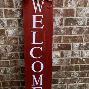 May include: A red wooden welcome sign with white lettering. The sign is decorated with a burlap bow and white and burgundy flowers.