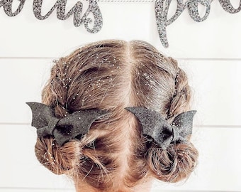 May include: A young girl with brown hair styled in two buns with black bat hair clips. The hair is decorated with glitter. The girl is wearing a white shirt and is facing away from the camera. A silver glitter banner with the words "Boo" and "Party" is hanging above the girl.