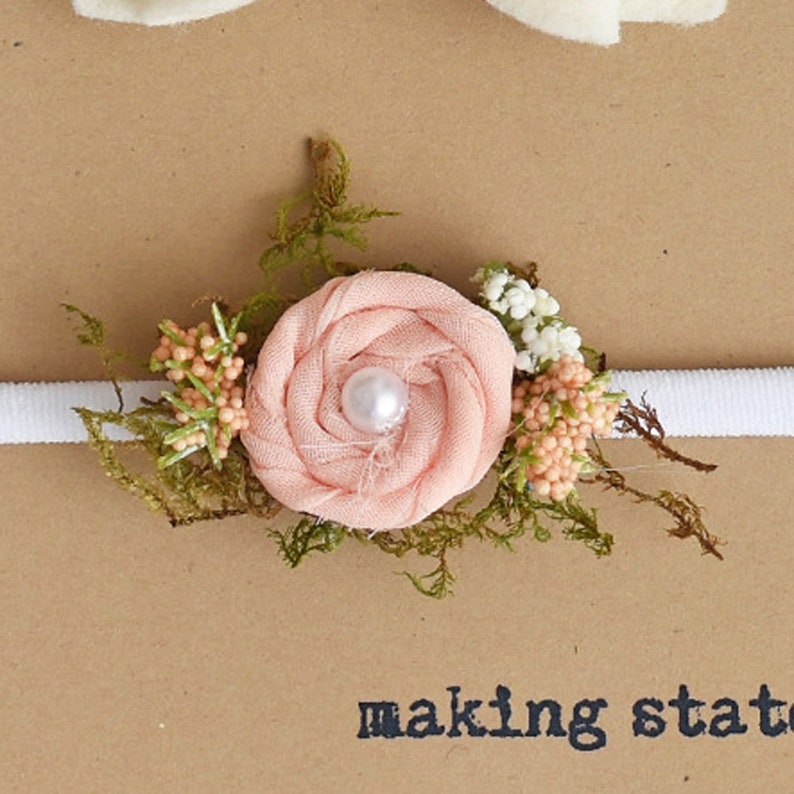 May include: A peach-colored fabric flower with a pearl center, surrounded by greenery and small white and peach-colored flowers, is attached to a white elastic headband.