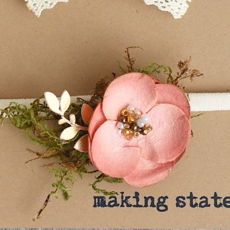 May include: A pink felt flower with gold accents and a cluster of beads in the center, attached to a white elastic headband. The headband is on a brown surface with the text "making state" printed on it.