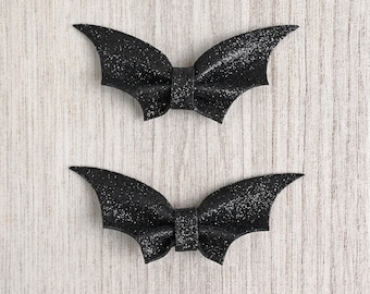 May include: Two black glitter bat-shaped hair clips with a bow in the center.