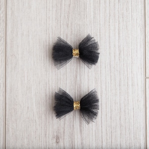 May include: Two black tulle bows with gold centers. The bows are on a white wooden background.