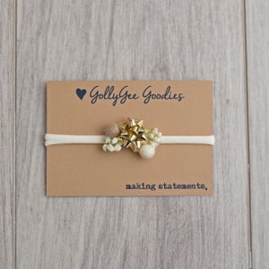 May include: A white headband with a gold bow, white flowers, and brown pom-poms. The headband is on a brown card with the text "Golly Gee Goodies making statements."