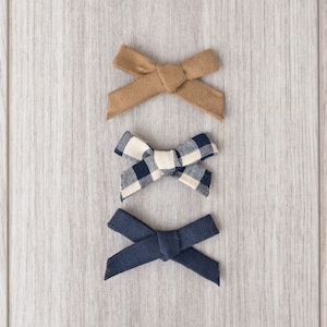 May include: Three fabric hair bows in different patterns. The top bow is brown, the middle bow is black and white checkered, and the bottom bow is navy blue.