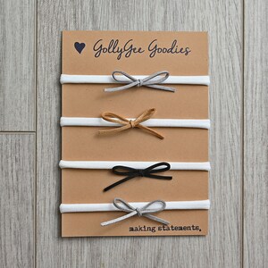 May include: Four white elastic headbands with different colored suede bows. The bows are gray, brown, black, and gray. The card reads "Golly Gee Goodies making statements."
