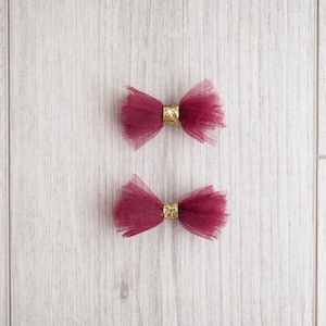May include: Two burgundy tulle bows with gold glitter centers. The bows are on a white wooden background.