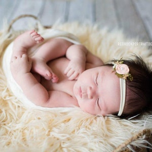 May include: A newborn baby girl sleeping peacefully in a white faux fur basket. She is wearing a white headband with a pink flower.