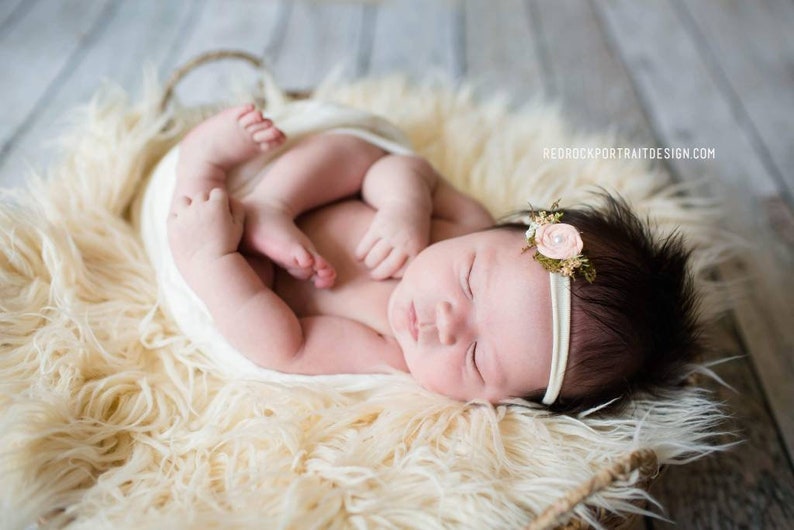 May include: A newborn baby girl sleeping peacefully in a white faux fur basket. She is wearing a white headband with a pink flower.