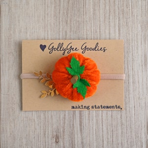 Peut inclure: Un serre-tête avec une citrouille orange en feutre et des accents de feuilles dorées. Le serre-tête est sur un fond brun avec le texte "Golly Gee Goodies making statements."
