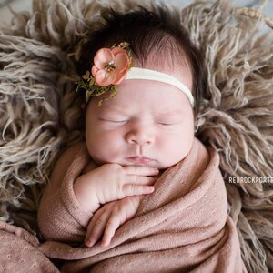 May include: A newborn baby girl is sleeping peacefully in a brown fuzzy basket. She is wearing a pink swaddle and a headband with a peach-colored flower.