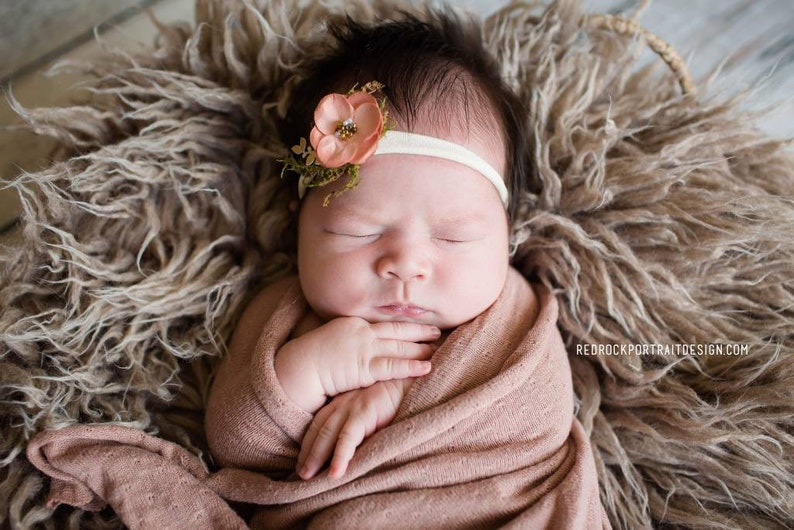 May include: A newborn baby girl is sleeping peacefully in a brown fuzzy basket. She is wearing a pink swaddle and a headband with a peach-colored flower.