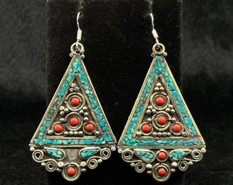 May include: A pair of silver-toned triangle earrings. Each earring features a turquoise mosaic design around the edges and red circular accents. The earrings have a decorative, ornate design and are displayed on a black surface.