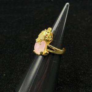 May include: Gold ring with a pink stone and a detailed frog design. The frog has a red stone for an eye.