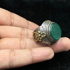 May include: A silver ring with an oval green stone and intricate gold detailing. The ring is set with a large, oval-shaped green stone, surrounded by a silver band with intricate gold detailing.