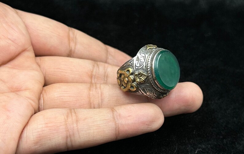 May include: A silver ring with an oval green stone and intricate gold detailing. The ring is set with a large, oval-shaped green stone, surrounded by a silver band with intricate gold detailing.
