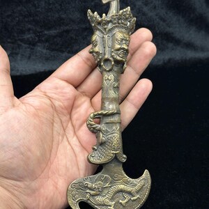 May include: A bronze-colored, ornate ceremonial dagger. The handle features two faces, intricate carvings, and a dragon design on the blade. The dagger has a curved blade and a horse-shaped finial at the top. The overall design is detailed and artistic.