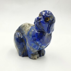 Op de afbeelding: Een klein, gesneden lapis lazuli beeldje van een hond die zit met zijn kop omhoog gekanteld. Het beeldje is diepblauw met witte en gouden accenten.