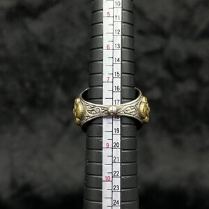 May include: A silver and gold ring with intricate designs and a small pearl accent. The ring is measured against a ruler, showing a size of 18.