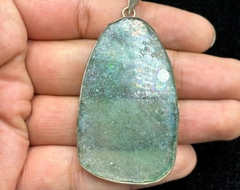 Pode incluir: Um pingente com uma argola prateada e uma pedra grande, de formato irregular, verde e azul. A pedra tem uma superfície texturizada e iridescente, com uma borda de metal prateado. O pingente está na mão de uma pessoa.