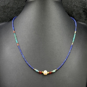 May include: A delicate necklace featuring small, dark blue beads with accents of turquoise, red, and gold. The focal point is a large, creamy white pearl. The necklace is displayed on a black mannequin.