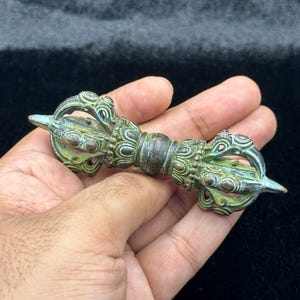 May include: A bronze-colored, ornate vajra, a ritual object, is held in a hand. The vajra features intricate designs and a central handle. The object has a weathered appearance with hints of green and blue, suggesting age and use. The background is a dark, solid color.